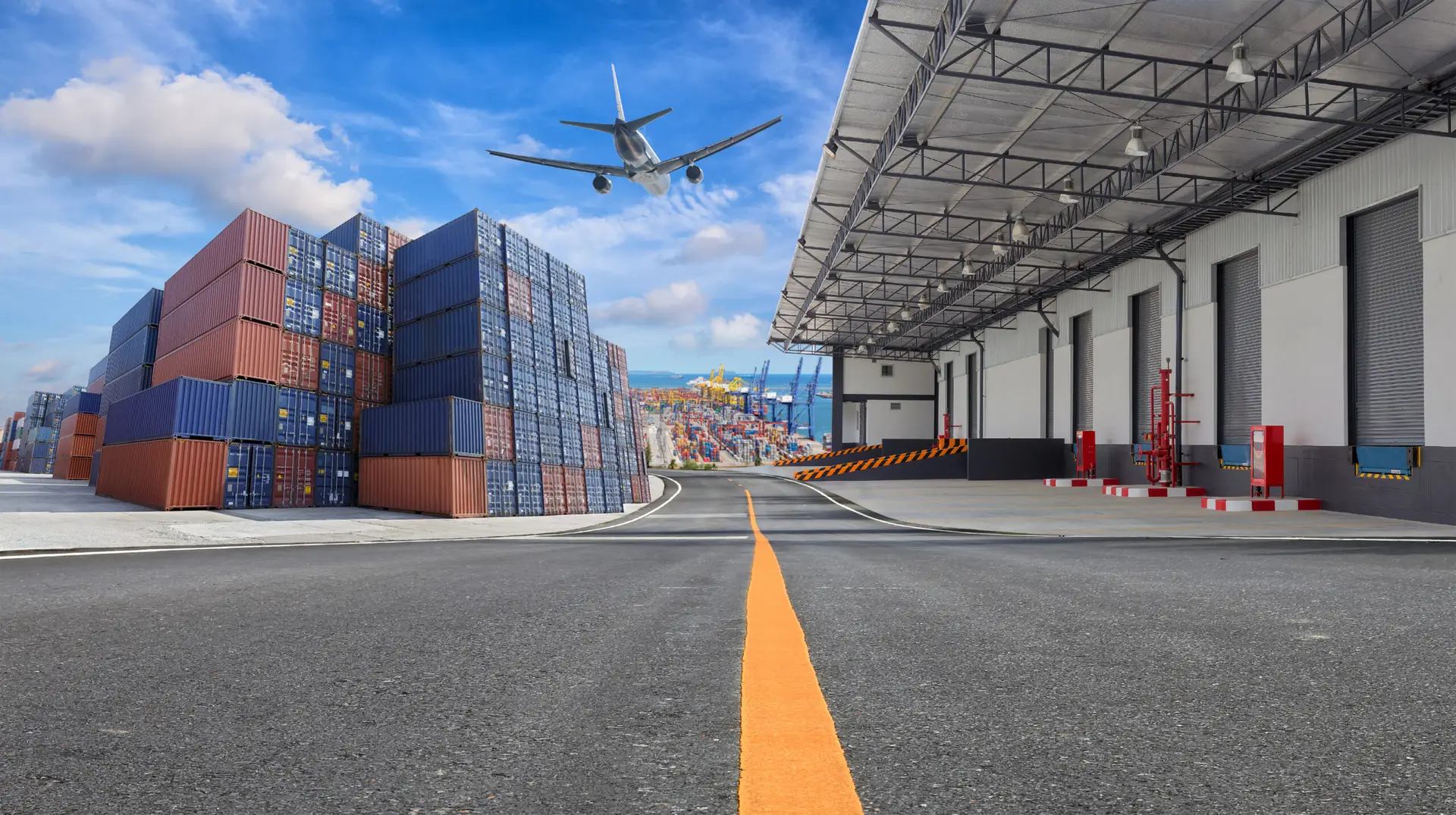 Shipping containers stacked beside a warehouse with an airplane overhead at a seaport.